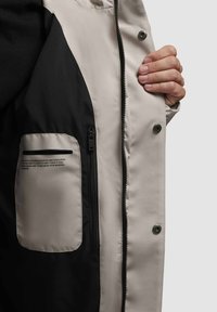 Chaqueta de color claro con un interior negro, que presenta un bolsillo con cremallera y botones a presión. La tela parece suave con un acabado mate.