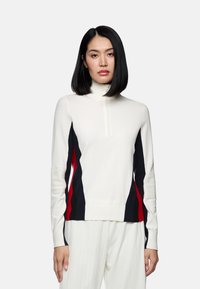 Weißer Pullover mit langen Ärmeln und hohem Kragen, der an den Seiten rote und marineblaue diagonale Streifen aufweist, eine gerippte Textur an den Ärmeln hat und mit einem Reißverschluss ausgestattet ist.