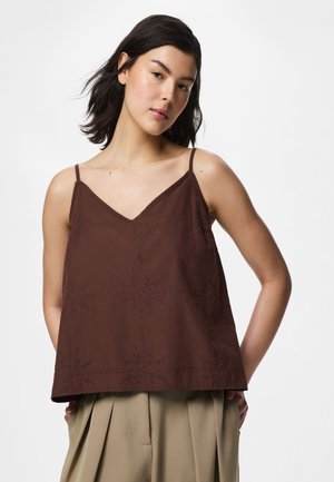 Kvinde med mørkt hår iført en ærmeløs brun broderet top og beige plisserede bukser, poserer foran en ensfarvet hvid baggrund.