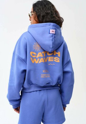 Osoba nosząca niebieski zestaw z kapturem i szortami z "Catch Waves" oraz pomarańczową grafiką słońca nadrukowaną na plecach.