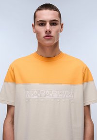 Kortærmet T-shirt med orange øverste sektion og beige nederste sektion. Har et præget logo "NAPAPIJRI geographic" i hvid på forsiden.