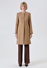 Cappotto di lana cammello con un design dritto, colletto rotondo e tre bottoni. Abbinato a pantaloni marroni a vita alta e scarpe a punta.