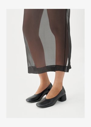 Scarpe con tacco a blocco nero in pelle liscia, indossate con una gonna nera trasparente con orlo pulito e design minimalista.