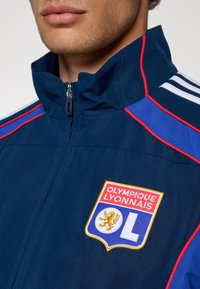 Marineblaue Sportjacke mit weißen und roten Akzenten, die mit einem gestickten Olympique Lyonnais-Logo, hohen Kragen und Reißverschluss ausgestattet ist.
