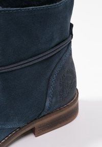 Niebieski zamszowy botek za kostkę z okrągłym noskiem, teksturowanym materiałem, dekoracyjnym przeszyciem oraz drewnianym obcasem z grawerowanymi detalami.