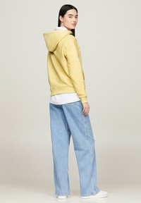 Felpa gialla con cappuccio e vestibilità rilassata, abbinata a jeans a gamba larga blu chiaro. Texture liscia, design semplice e accenti minimi.