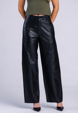 Vrouw draagt zwarte leren broek met hoge taille en wijde pijpen, gecombineerd met een groen cropped topje en zwarte pumps met puntige neus.