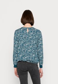 Turkos blus med blommönster, rund halsringning, nyckelhålsdetalj på baksidan och långa ärmar med kragade manschetter. Strukturerad tyg.