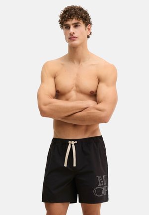 Man zonder shirt met krullend haar, armen gekruist, draagt zwarte zwemshorts met witte trekkoord en letters op het linkerbeen.