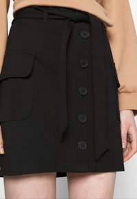 Jupe en tissu noir avec ceinture, boutons sur le devant et poches latérales. Texture lisse avec une coupe droite et un détail minimaliste.