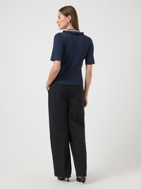 Femme portant un haut en maille côtelée bleu marine à manches courtes avec un col à volants et un pantalon noir à jambes larges, se tenant les mains dans les poches devant un fond blanc.