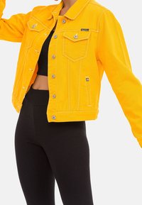 Yellow denim jacket with a cropped design, two chest pockets, silver hardware, and button closures. Paired with black ribbed leggings.