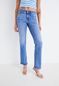 Jeans de mezclilla azul claro con diseño de pierna recta, que cuentan con cinco bolsillos y un cierre de boton. Llevados con sandalias de tacón transparente.