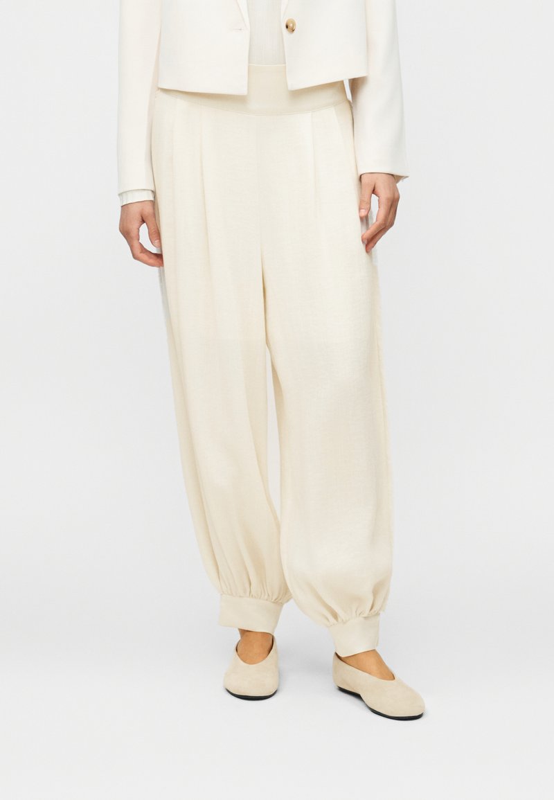 Άτομο που φοράει φαρδιά εκρού παντελόνια με μαζεμένες μανσέτες, ταιριαστό σακάκι και μπεζ επίπεδα παπούτσια, στέκεται μπροστά σε απλό φόντο.