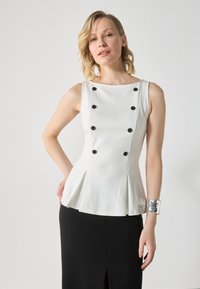 Vit peplumtop med svarta knappar, ärmlös design, slät yta och subtila veck, kombinerad med en svart kjol och ett silverfärgat armband.
