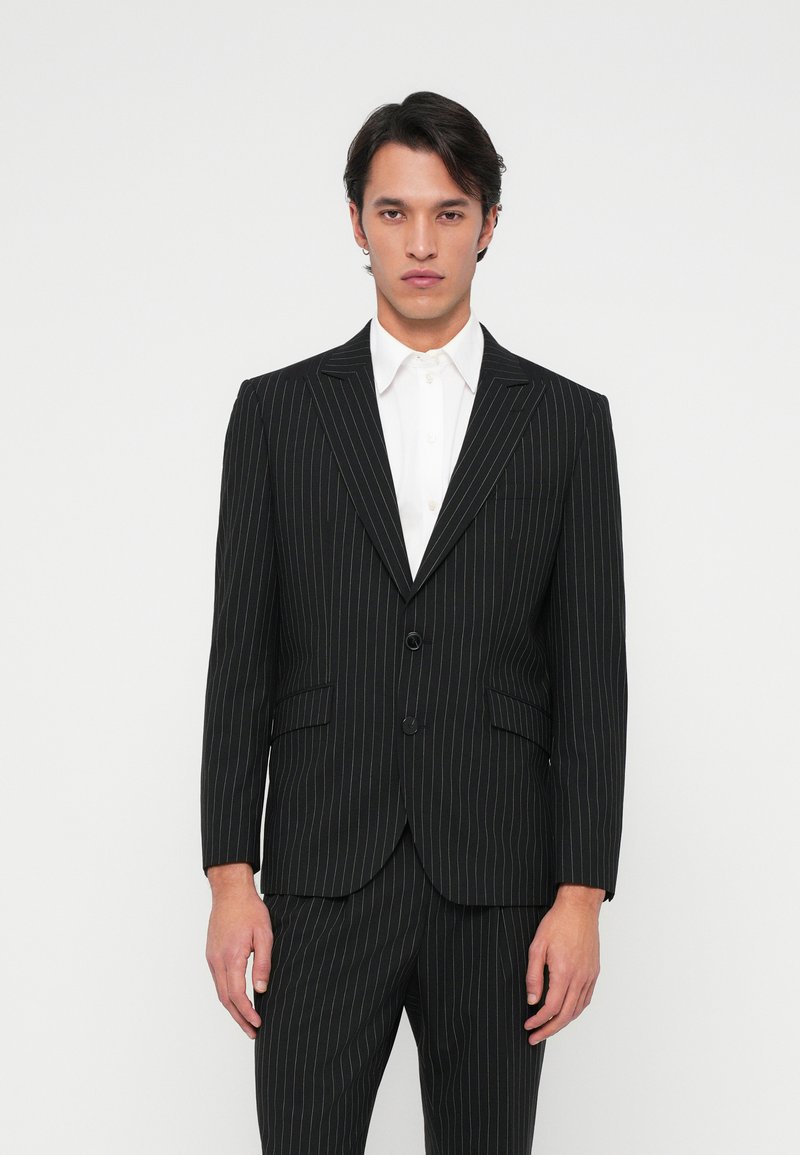 Μαύρη σακάκι με ρίγες και πέτο με βαθούλωμα, δύο κουμπιά και δύο μπροστινές τσέπες. Φοριέται πάνω από ένα λευκό πουκάμισο με γιακά.