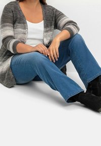 Cardigan de punto gris y blanco con diseño a rayas, combinado con jeans acampanados azules y botas negras hasta el tobillo. Posición sentada, fondo neutro.
