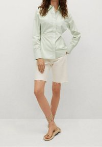 Hellgrübes Langarmhemd mit Knöpfen, kombiniert mit elfenbeinfarbenen fransigen Shorts und beigen sandaletten mit Riemen. Glatter Stoff mit taillierter Passform.