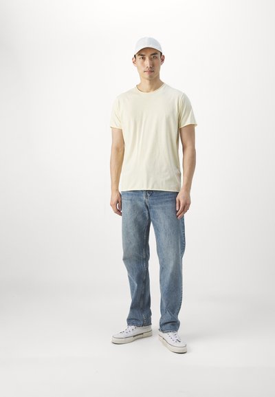 T-shirt jaune clair à manches courtes, jeans bleus et baskets blanches ; la personne se tient debout avec une casquette grise clair. Tenue simple et décontractée.