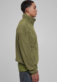 Pullover in pile verde oliva con colletto alto, frontale con zip e maniche lunghe. Caratterizzato da una vestibilità rilassata e un tessuto testurizzato.