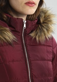 Bordeauxfarvet dunjakke med lynlås og hætte foret med imiteret pels. Markerede sømme tilføjer tekstur og struktur. Sølvfarvet lynlåsdetalje.