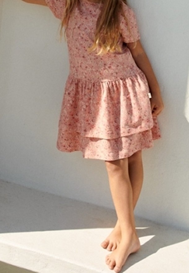 Enfant debout pieds nus sur un rebord blanc, portant une robe rose à fleurs à manches courtes avec une jupe superposée, devant un mur clair uni.