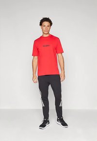 Raudonas medvilninis marškinėlis su juodu Adidas logotipu, derinamas su juodomis kelnėmis su baltomis juostelėmis šonuose ir juodais sportbačiais.