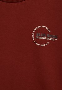 Bordeauxtröja med mjuk textur, prydd med cirkulär vit och grå text: "BLUE RIDGE" tillsammans med motiverande fraser. Klassisk crew neck-design.