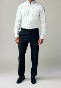 Chemise blanche à boutons, associée à un pantalon sombre bien ajusté, une ceinture noire et des chaussures de ville noires brillantes, formant un ensemble formel.