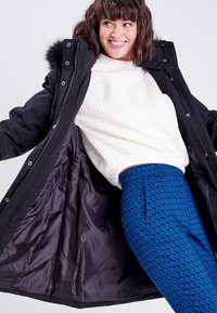 Parka d'hiver noire avec capuche en fourrure, ouverte pour révéler un intérieur brillant. Associée à un pull texturé blanc et à un pantalon bleu à motifs.