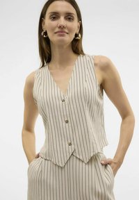 Chaleco sin mangas beige con rayas verticales negras, cuello en V y dobladillo puntiagudo. Presenta cuatro botones en la parte delantera; textura de tela suave.