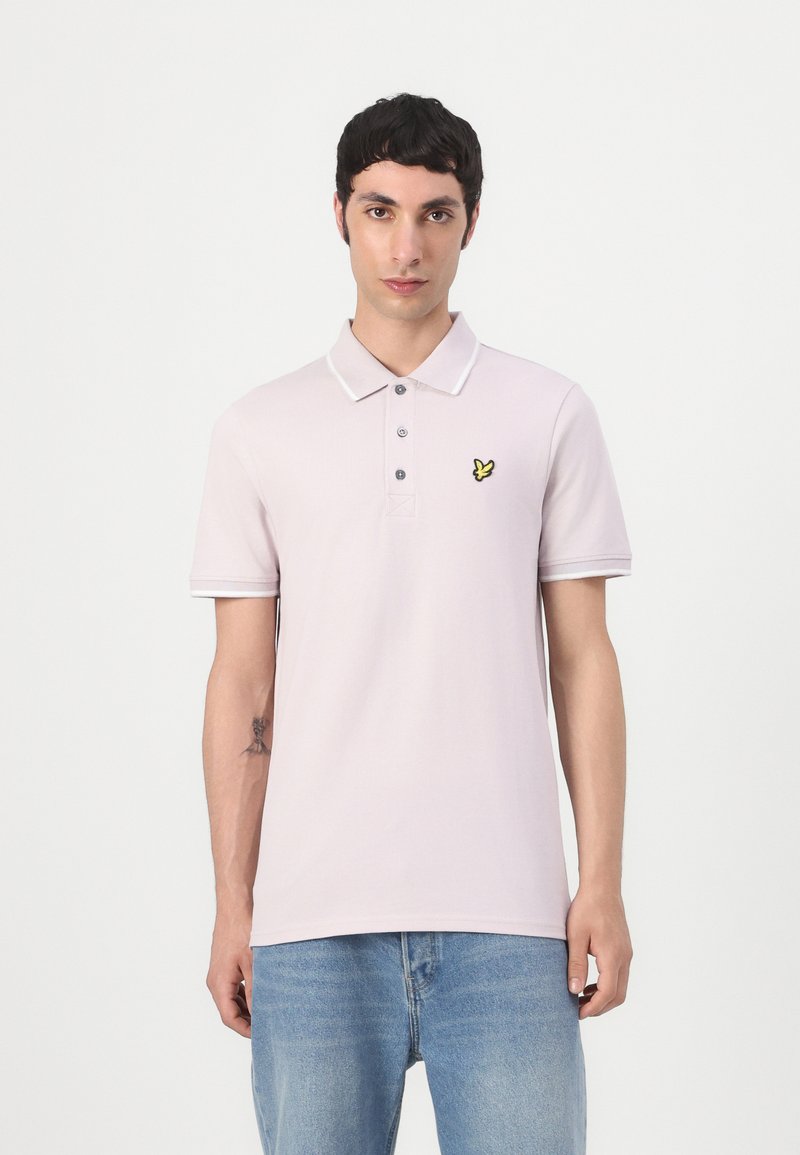 Jeune homme aux cheveux courts et foncés portant un polo rose clair avec un logo d'oiseau jaune et un jean bleu, debout sur un fond blanc uni.