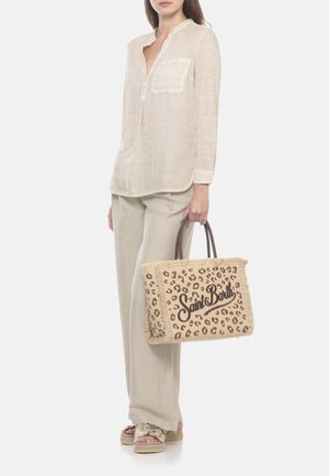 Femme portant une chemise et un pantalon beige, tenant un sac cabas tissé avec imprimé léopard et texte "Saint Barth", debout sur un fond blanc.