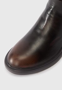 Bottine en cuir marron avec une texture lisse, bout légèrement arrondi et semelle en caoutchouc noir. Détails de couture visibles sur le dessus.