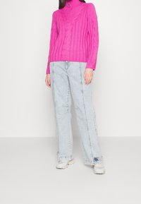 Pull à col roulé en maille fuchsia avec texture côtelée, associé à un jean bleu clair à coupe ample avec des détails de couture subtils et des coutures latérales.