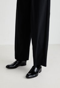 Pantofi de piele negri, cu un finisaj lucios, având un vârf neted și un toc jos, asortați cu pantaloni negri lejera.