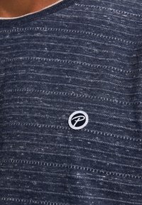 Tricou din material bleu marin, cu dungi texturate orizontale, având guler rotund și o inscripție circulară albă pe piept.