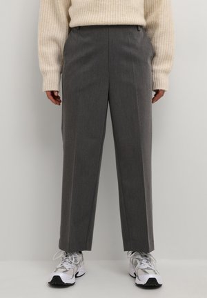 Personne portant un pantalon gris à jambes larges, un pull en maille blanc et des baskets blanches, debout devant un fond uni.