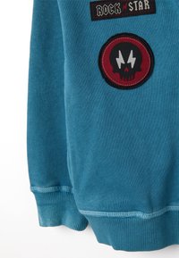 Sudadera azul con un parche de "ROCK n' STAR" y un parche circular rojo con diseño de calavera. Puños acanalados y textura suave.