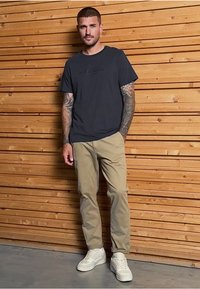 Tmavě šedé bavlněné tričko s logem, khaki chinos a bílé tenisky. Osoba má viditelné tetování a stojí u dřevěné zdi.