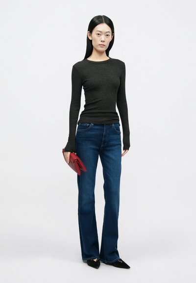 Γυναίκα με ίσια μαύρα μαλλιά, φορώντας μαύρο μπλουζάκι με μακριά μανίκια, μπλε τζιν, μαύρα μυτερά παπούτσια, κρατώντας κόκκινη τσάντα φάκελο σε λευκό φόντο.