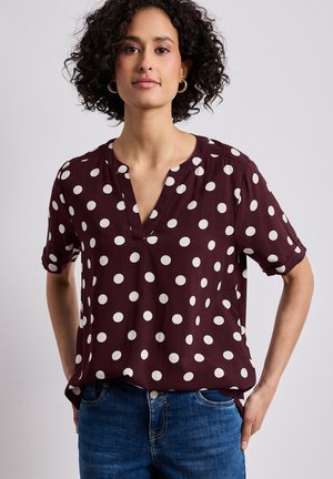 Frau mit lockigem Haar, die goldene Creolen, eine weinrote kurzärmelige Bluse mit weißen Punkten und blaue Jeans trägt, steht mit den Händen in der Hüfte.