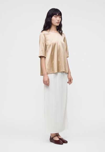 Femme aux longs cheveux noirs portant un haut beige brillant à manches courtes, une jupe plissée blanche longueur cheville et des ballerines marron foncé, debout de profil.