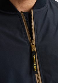 Chaqueta de bombardero navy con cremallera dorada y cuello de canalé negro. Presenta un tirador de marca con "PME LEGEND" en amarillo en la cremallera.