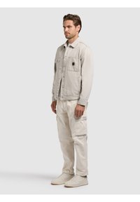 Homme debout portant une veste décontractée beige clair, un pantalon cargo assorti et des baskets blanc cassé sur un fond uni.