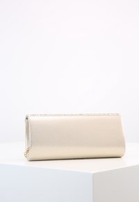 Rektangulær metallisk guld clutch taske med en tekstureret topkant, der hviler på en hvid kube mod en neutral baggrund.