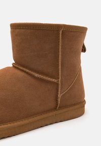 Tan ankle boot made of soft suede, featuring a rounded toe, stitched seams, and a textured collar. The sole is flat and sturdy.