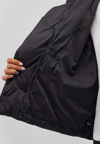 Giacca nera con texture liscia e lucida, design di lunghezza intermedia, chiusura con zip e coulisse regolabile all'orlo.