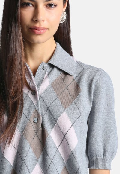 Femme aux longs cheveux foncés portant un pull gris boutonné avec un motif losanges rose et marron et une grande boucle d’oreille ronde en argent.
