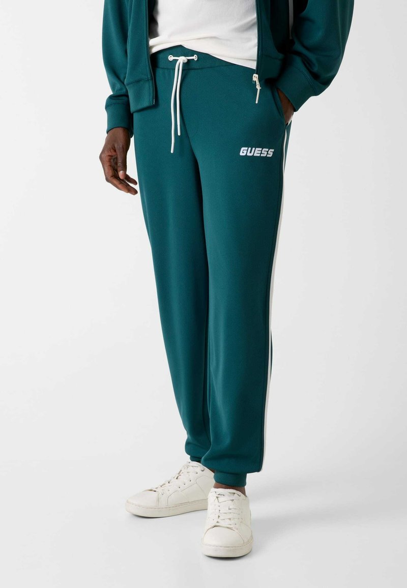 Turkooizen trainingsbroek met een witte trekkoord en het GUESS-logo, met een gestructureerde stof en zijstrepen. Draagbaar met witte sneakers.